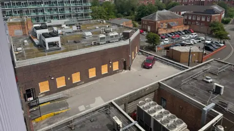 Annabel Amos/BBC Aerial shot showing roofs of commercial buildings, some with lit-up windows. There is also air conditioning / heating plant. There is a red car parked on one of the levels of roof.