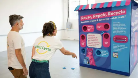 East London Waste Authority A large pink and blue cube carrying slogans about battery recycling. A man a woman are throwing items at it, presumably in a game.
