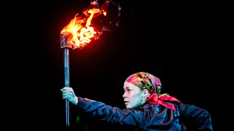 A young performer on stage holds aloft a fire lit torch against a dark backdrop.. They wear a colourful bandana on their head.
