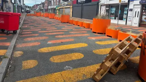 A street painted with orange and yellow blobs right across the pavement and carriageway. There are orange tubs and planters filled with dead plants. In the foreground you can see an abandoned wooden pallet. 