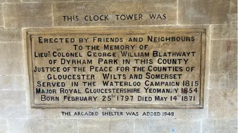 A stone plaque gives details of George William Blathwayt - who the Chipping Sodbury clocktower celebrates. The carved letters are painted black to highlight them against the lighter Cotswold Stone surround.