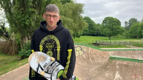 A man wearing a black hoodie with yellow detailing is holding a skateboard and is stood at the top of a skateboard park. There are fields and trees in the distance.