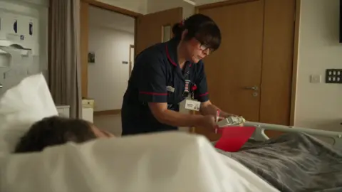 Image of a care staff member at a hospice bed which has a patient in it. She is wearing blue overalls and is looking at medication.