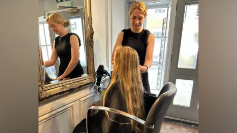 Alex Chapman Nine-year-old Noah sitting in a hairdressers as he prepares to get his long blond hair cut for charity. The hairdresser stands on his right hand side preparing to cut his hair.