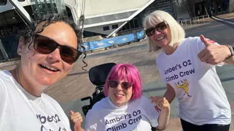 A selfie of Amber Donovan, Kayleigh Jennings and another woman outside the 02 Arena. Ms Donovan has pink hair in the style of a bob and wears sunglasses. Ms Jennings has black curly hair and wears sunglasses. They all wear white T-shirts that read Amber's 02 Climb Crew.