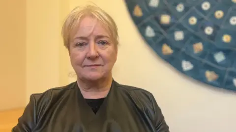 Karen sits in an office with light‑coloured walls. Behind them is a large circular decorative wall piece featuring patterns and symbols. A wooden desk is partially visible to one side.