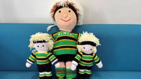 Carroll Weston/BBC Three knitted versions of Henry Pollock, wearing the Saint rugby kit, with black, yellow and green strips. They all have blonde hair and a black hairband. They are placed on a blue sofa. 