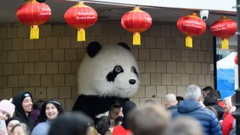 Birmingham Hippodrome A large panda mascot stands beneath rows of red Chinese lanterns as crowds gather around it. The panda has a round white head with black markings and is positioned in a sheltered outdoor area. People in coats and jackets fill the foreground.
