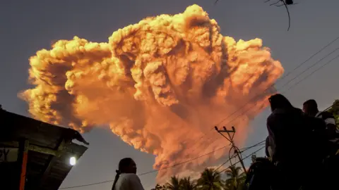 A vast orange plume erupts into the sky above several people standing in the foreground.