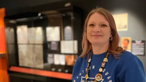 Emily Shepperson smiles at the camera as she stands in front of a glass display case that holds letters. She has light-brown hair and is wearing a blue jumper with a lanyard around her neck. 