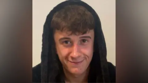 Shay Bladen smiling into the camera. He has brown, short hair and has a navy blue dressing gown hood over his head.