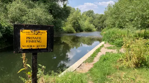 A river is pictured and trees alongside it. There is a ledge and beside it is a sign which says "private fishing"