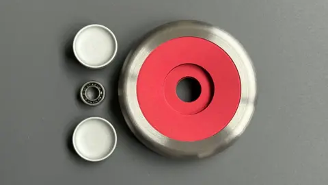 James Goh A fidget spinner is placed on a table. It has been taken apart, revealing its components. The central spinner has a red core with a silver outer rim. Beside it are three separate metal pieces laid out neatly, likely parts of the device. The setup highlights the construction and materials used, with a focus on the engineering detail of the spinner.