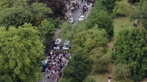 Essex Police A drone image of two large groups of people being held back by a police cordon. They are standing in a road which is lined by trees and greenery.