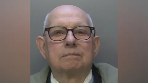 A bald man with short grey hair around the side of his head. He has black glasses.