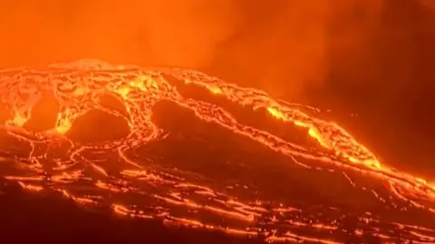 Lava spills down the side of a volcano, as an orange haze glows in the background