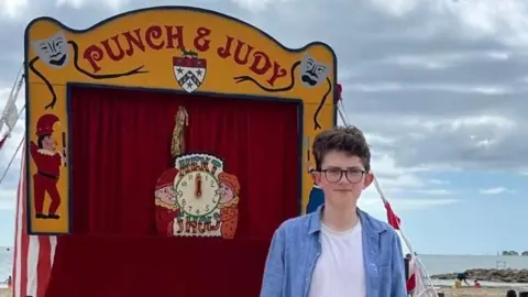Family photo William Vaughan posing for a picture in front of a Punch and Judy tent at the beach. People are seen walking behind him in the distance. It is a cloudy day.