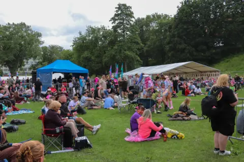 BBC / JOSH PARRY Crowd gathered on a grassy field with tents and flags, sitting and socialising at an outdoor community event