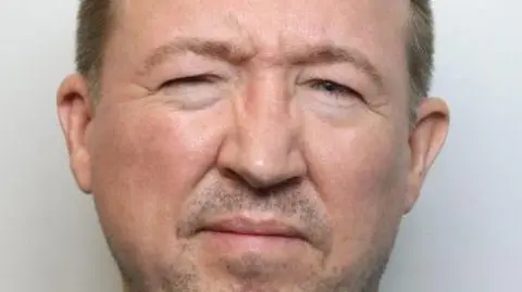A man is looking at a camera for a police mugshot, squinting his eyes