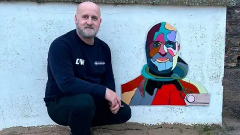 Darren Wiles Darren Wiles crouched next to a multi-coloured mural of his face on a white wall