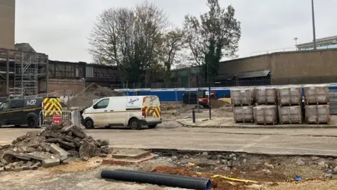 LDRS Two small vans with high visibility patterns on them are parked on a concrete area. Pallets piled with construction materials are on the right of the vans. An area in front of them has been dug up and there is a pile of concrete. In the background are two trees, a wall and a building. 