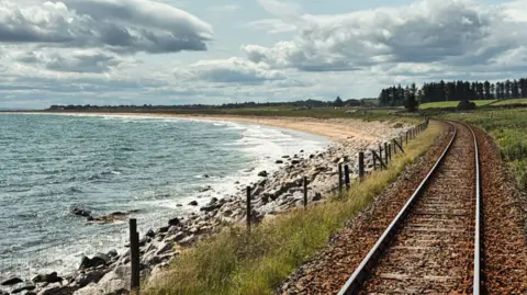 Train tracks running alongside the coast, with a mixture of rocks and sand on the beach and trees in the background.