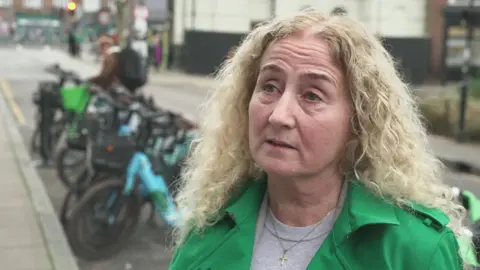 A woman with curly blonde hair wearing a green jacket, standing on the pavement in a residential street. There is a row of parked e-bikes in the background behind her.
