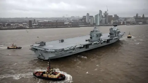 Royal Navy HMS Prince of Wales on the River Mersey, with the Liverpool city skyline in the background showing the tall buildings