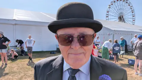 Isaac Chenery/BBC A man wearing tinted glasses smiles at the camera. He is wearing a black bowler hat, suit jacket, blue shirt and black tie. Behind him are white marquees and a Ferris wheel. 