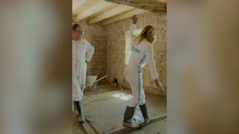 A man in white overalls looks on as a woman in white overalls walks along a narrow piece of wood over wet concrete