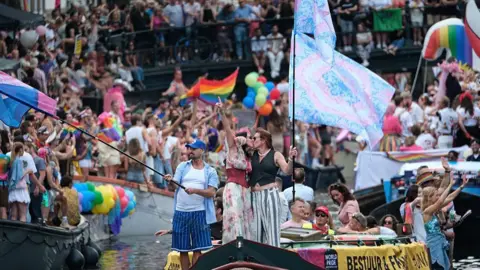 Getty Images Two people kiss on the deck of a boat decorated with "WorldPride" banners during a canal parade, surrounded by cheering crowds lining the canal, with flags including a large trans flag visible in the foreground.