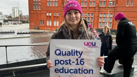 Carys Davies in pink UCU hat holding placard. She has long blonde hair and wears a brown puffer coat.