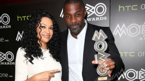 Kanya King and Idris Elba standing together, she's pointing at him and he's holding a Mobo trophy