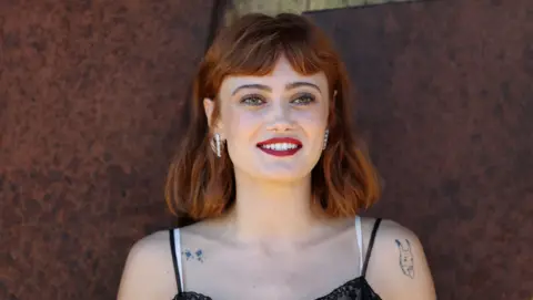 Getty Images Ella Purnell em frente a um fundo marrom. Ela tem tatuagens nos ombros, cabelos ruivos e brincos de prata. Ela está sorrindo.