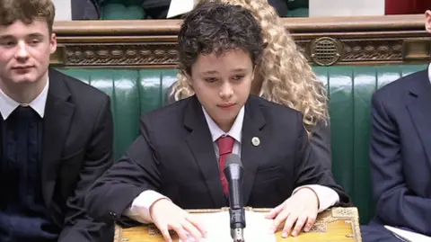 Owen Glass stands at the dispatch box. He is wearing a white shirt, dark red tie, and black blazer. He has brown curly hair.