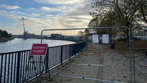 A footpath along the River Tees is blocked off by metal grating. A red sign reading "FOOTWAY CLOSED" is placed in front. A bridge of the river can be seen in the distance.