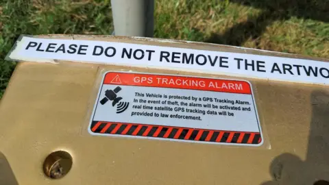 A close up on the gold toilet, showing a sign reading: This Vehicle is protected by a GPS tracking Alarm. In the event of theft the alarm will be activated and real time satellite GPS tracking data will be provided to law enforcemnt,