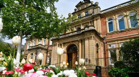 The exterior of Penrith Town Hall. It has an ornate stone, sand-coloured entrance porch which creates a balcony at the first floor. There are colourful flowers, trees and bushes in front of the red stone building.