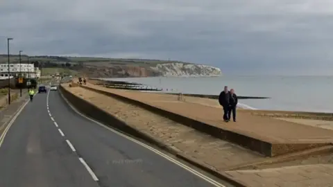 Google A Google image showing two people walking along the sea defences between a beach and a road with cliffs in the background. 