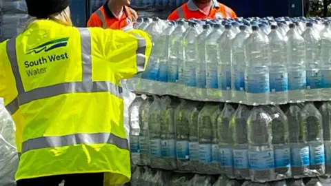 Bottled water stacked on a wooden pallet in front of a blue lorry. There is a person wearing a yellow hi-vis jacket, with the words 'South West Water' on the back. 