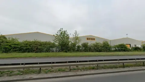 Google A long white warehouse is partially concealed behind a row of trees and shrubs but the word Bertschi is clearly visible on the side of the central part of the building 