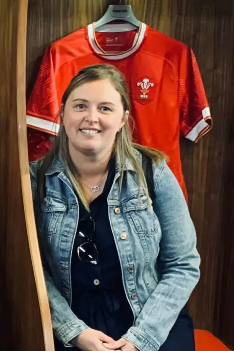 Emma Coomer A woman smiles at the camera in front of a rugby jersey. She has long straught brown hair and wears a denim jacket, black t-shirt and a silver necklace.