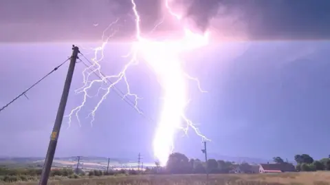 Johnny B A thick white lightning strike captured from across a field. Telephone poles and overhead wires stand in the foreground, with rolling hills in the background beneath a grey, stormy sky.