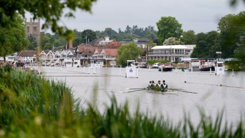 A crew of four people row a boat along a stretch of river, with buildings in the background and grass in soft focus in the foreground