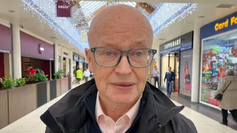 Peter is pictured standing in a shopping centre. He is wearing glasses, a pink shirt and a black coat 