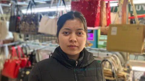Preet, an Asian woman with dark hair and brown eyes, standing in front of handbags on display on her stall.