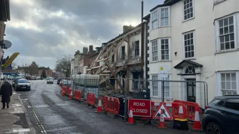 A fire damaged building being propped up by wooden beams. A white building next door is also cordoned off. The cordon takes up much of the road leaning to just one lane being usable
