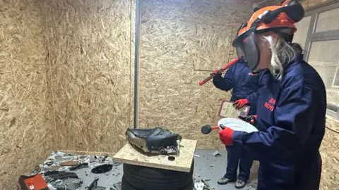 Tim Seward Claire Seward in the foreground dressed in mauve overalls and wearing an orange helmet and holding a mallet in a room covered in chipboard with a smashed up small appliance in the centre 