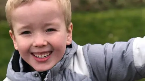 A boy with short blonde hair and a grey coat smiles as he holds out his left arm.