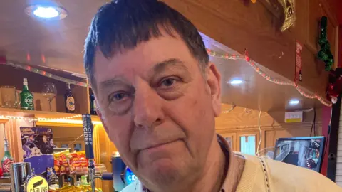 Sam Grainger is standing in a bar. There are alcohol taps behind him. He is looking into the camera and has a half-smile.  He has short dark hair and wears a yellow jumper over a faded pink shirt.